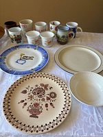 Photo of one Staffordshire Made in England decorative plate with floral pattern, two Corelle plates, one white disposable/plastic plate, and eight assorted ceramic mugs with various designs arranged on a tabletop.