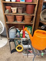 Wooden shelf containing terracotta pots, an orange plastic watering jug, a vintage metal watering can, assorted garden sprays and tools, and a bag of fertilizer below.