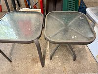 Photo showing two square outdoor side tables with textured tempered glass tops and metal frames resting on concrete floor.