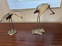 Wide view of both brass crane sculptures on a wooden surface, one larger on textured base, one smaller on round base