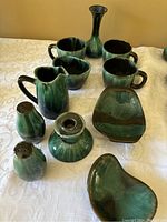 Green glazed pottery set arranged on a fabric surface showing the pitcher, mugs, vase, shakers, candlestick holder, and serving dishes.