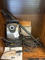 Stanley electric pressure washer with coiled power cord, attached hose and wand, small cleaning brush, and owner's manual set on wooden shelf.