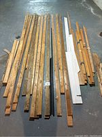 Wide view of assorted lengths of hardwood floor pieces and baseboard trim laid on concrete floor, showing natural wood and white painted pieces in various lengths and conditions.