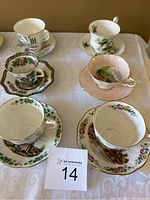 Set of five teacups and saucers displayed on a white lace tablecloth showing various floral and scenic patterns and different colors, including white, pink, and green bases with gold trim.