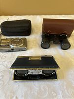 Sony Cyber-shot camera with black carrying case paired with two different vintage binoculars and cases on cream fabric.