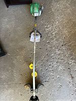 Full length view of the green gas powered weed eater laid flat showing engine, shaft and trimming head with guard.