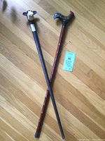 Two carved wooden walking sticks laid on wooden floor, one with cow head handle and one with rooster handle, both about 37 inches long.