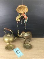 Front view of brass dishes, brass teapot with wooden handle, brass elephant figurine, and hanging elephant mobile against dark background.