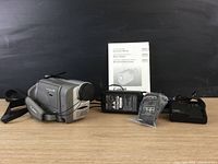 Canon ES8100V video camcorder lying on a table alongside remote control, battery charger, power adapter, and instruction manual on a black background.