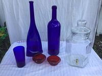 Photo showing two cobalt blue bottles of varying heights, small cobalt blue glass, two ruby red bowls, and clear glass beverage dispenser with spigot, all arranged on white surface with cloth backdrop.