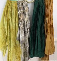 Four scarves hanging including a yellow lightweight scarf, a beige plaid silk scarf with fringe, a deep green silk scarf, and a mustard yellow silk scarf all appearing lightweight and gently used.