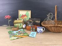 Photo showing multiple vintage and antique decorative items arranged on a wooden surface including framed embroidered artwork, small metal storage box, ceramic pitcher, wire basket, iron, floral metal box, decorative rose, salt and pepper shakers, and two embroidered cloth pieces.