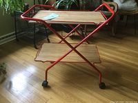 View of the full bar cart showing X-shaped red metal frame, upper shelf with wood tone, and plastic casters.