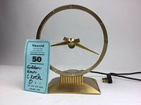 Front view of the golden hour clock showing the brass base, circular frame with hour markers, broad brass hands, and attached power cord.