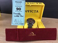 Invicta quartz watch in black stainless steel with orange accents resting on pillow inside yellow Invicta box, red Jordache soft watch case nearby.
