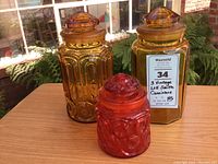Three vintage glass cannisters including two amber and one flame colored, with matching lids, displayed on wood table with outdoor vegetation background.