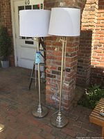 Pair of silver tone metal floor lamps shown side by side, full height, with white fabric shades. Both lamps stand 58.5 inches tall with round stepped bases and slender stems.