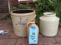Set of three unmarked pottery pieces: two large crocks and one jug on a brick floor with plants in background.