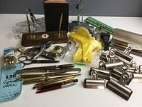 Overview of vintage office supplies including pens, binder clips, bookends, scissors, letter openers, and other metal tools on a white surface.