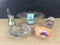 Five assorted glass items on wood surface against black background including a large clear ruffled edge bowl, a multifaceted clear crystal bowl, two rounded floral paperweights, and an orange-purple swirled glass dish.