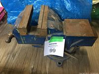 Photo of metal bench vice on wooden surface with blue tarp background. Shows jaws, handle, and mounting base.