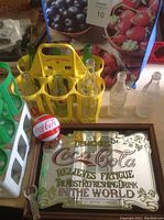 Photo of yellow plastic Coca-Cola bottle carrier with six vintage glass bottles and five loose clear bottles, plus Coca-Cola mirror sign below