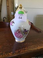 Front view of decorative urn showing hand-painted figures and floral designs with gold accents and finial on lid