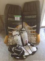 Photo showing two large Simmons goalie leg pads standing upright with four Bauer shin guards arranged in front and one Bauer shoulder pad at bottom.