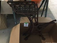 Pair of old rusted cast iron desk legs with decorative designs and chair support lattice, showing two legs positioned upright on cardboard in a workshop setting.