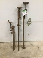 Three large old rusted metal bar clamps against a white wall, showing overall length and construction.