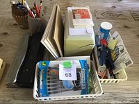 Overview of various stationery and office items arranged on a wooden table showing pens, papers, baskets, hole puncher.