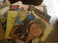 Stack of vintage vinyl record albums featuring John Denver and multiple other artists, showing wear consistent with age, arranged loosely on a surface.
