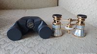 Bushnell 8x23 binoculars next to mother of pearl opera glasses on a light patterned fabric surface.