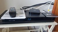Samsung Blu-ray player with remote and Apple TV device on top, cables in background.