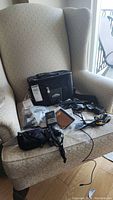 Sony Cyber Shot camera accessories arranged on a beige upholstered armchair, including carrying case, cords, and charger. Taken to show overall lot contents and condition.