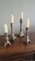 Four metal candle holders of varying heights with slightly used cream taper candles placed on a wooden table against a plain wall.