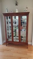 Wooden china cabinet shown from the front with three glass-panel doors displaying glassware inside. Decorative figurines are on top of the cabinet.