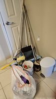 Cleaning supplies including mops, brooms, dustpan, bucket, and wastebasket placed on a tiled floor near a door.