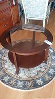 Photo showing round wooden coffee table with smoked glass center insert on top, lower wooden shelf, and four wooden legs. Table is placed on a patterned round rug.