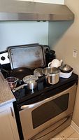 Cooking and baking items displayed on and near an oven including pots with lids, frying pan, cooking sheet or sieve, measuring cup, and a teapot.