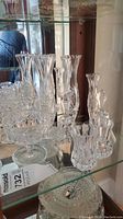 Crystal bud vases, dessert glasses, bells, pedestal bowl, and lidded dish arranged on glass shelf with reflective background.