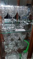 Fifteen crystal wine glasses on glass shelves arranged by pattern. Top shelf shows six glasses with geometric square-cut design. Lower shelf has six glasses with starburst cut and three plain crystal glasses. Clear stems and bases.