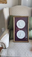 Framed set of two round white ceramic plates featuring angels, shown resting against a chair with green pillows, in a gold-tone frame with burgundy background.