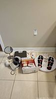 Photo of all health-related items including circulation promoter foot mat, back relief belt box, blood pressure monitor, mirror, hair dryer, compression nylons, and scale on tile floor