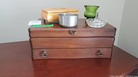 Photo showing a large wooden jewelry box with two drawers and metal pulls, a light brown rectangular box, a round silver metal container, a small translucent green glass container, and a clear cut-glass ring holder.