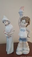 Photo of two Lladro porcelain figurines: a lady in bonnet and dress holding a rooster, and a boy with curly hair holding an Olympic flame torch.