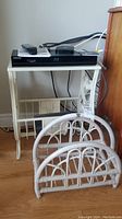 Small white metal frame table with clear glass top holding electronic items (remote, box). Two white wicker magazine racks sit on the floor in front of the table.