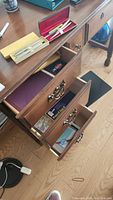 Wooden desk with three drawers open showing pens, pens boxes, binders, photo paper, and paper supplies in the drawers and on top.