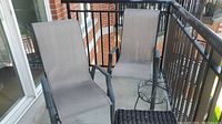 Two beige mesh fabric patio chairs with dark metal frames, a small woven dark brown side table, and a black metal plant stand on a balcony.