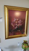 Full view of large framed print showing flowers in a vase on a piece of furniture, set on a wall above a stained glass decorative piece.